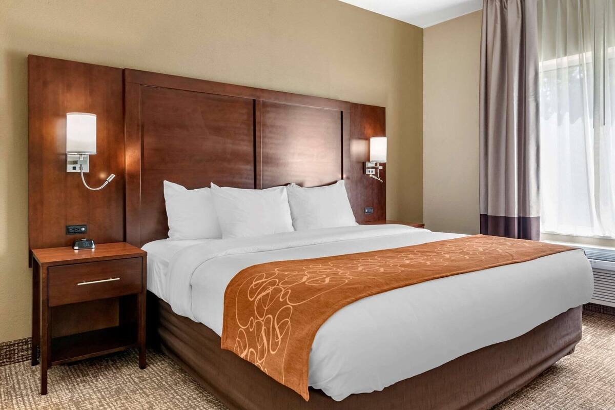 A hotel room with a large bed, a wooden headboard, bedside lamps, a nightstand, and a window with curtains.