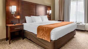 Habitación de hotel con una cama grande, cabecero de madera, dos mesitas de noche con lámparas, una mesita auxiliar y una ventana con cortinas.