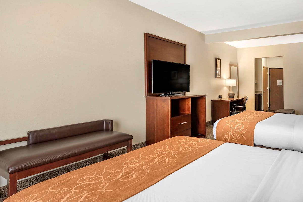 Habitación de hotel con dos camas, televisión de pantalla plana, banca, escritorio y espejo.