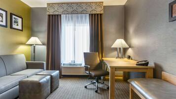 A hotel room with a sofa, a desk, a chair, a table, and a bench. There is a window with curtains and a wall-mounted air conditioner.