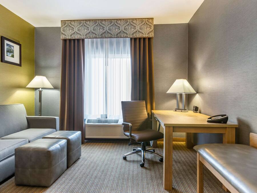 A hotel room with a sofa, a desk, a chair, a table, and a bench. There is a window with curtains and a wall-mounted air conditioner.