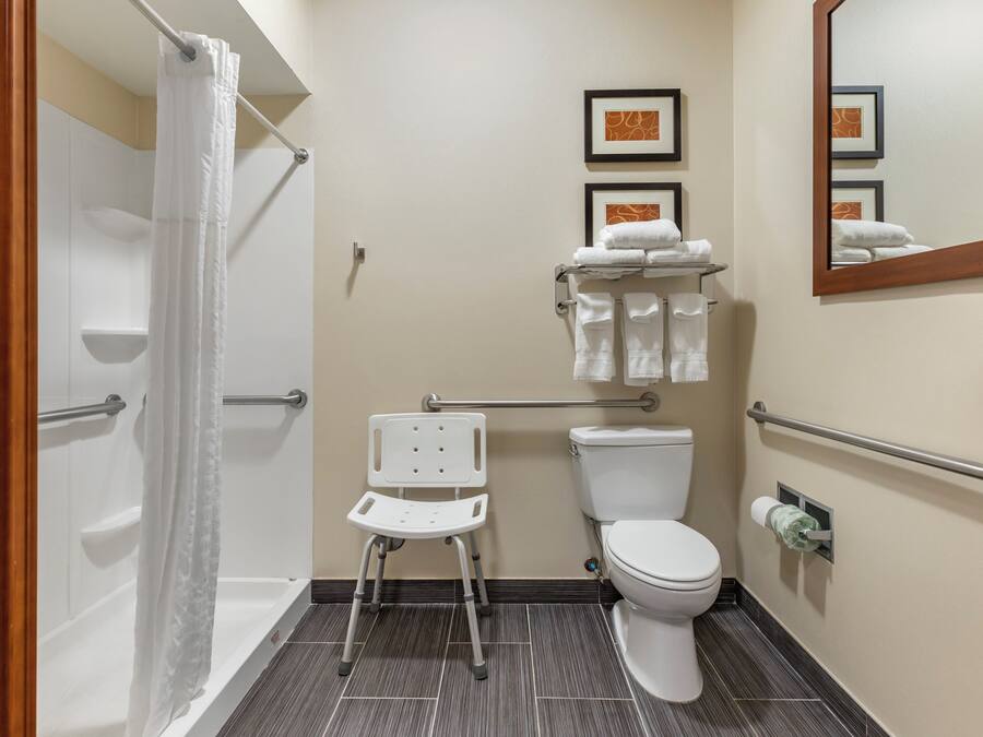 A bathroom with a shower, toilet, and a white chair. There are framed pictures on the wall and a mirror above the toilet.