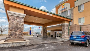 A Comfort Suites hotel entrance with a stone pillar, a covered driveway, and a blue SUV parked outside.