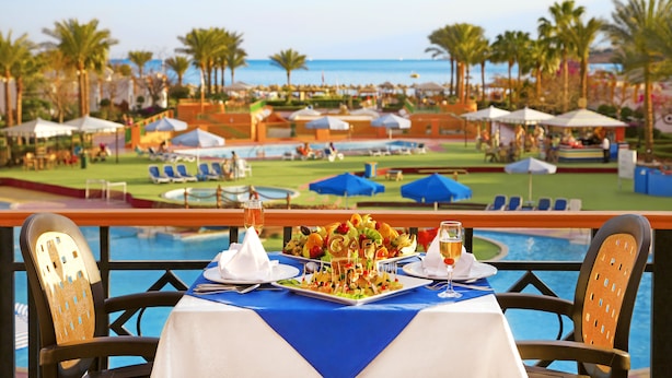 A dining table with a blue tablecloth, white plates, and a salad centerpiece, overlooking a pool and a beach area with umbrellas and lounge chairs. - Gafy Resort Aqua Park (Sharm El Sheikh)