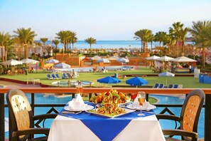 A dining table with a blue tablecloth, white plates, and a salad centerpiece, overlooking a pool and a beach area with umbrellas and lounge chairs. - Gafy Resort Aqua Park (Sharm El Sheikh)