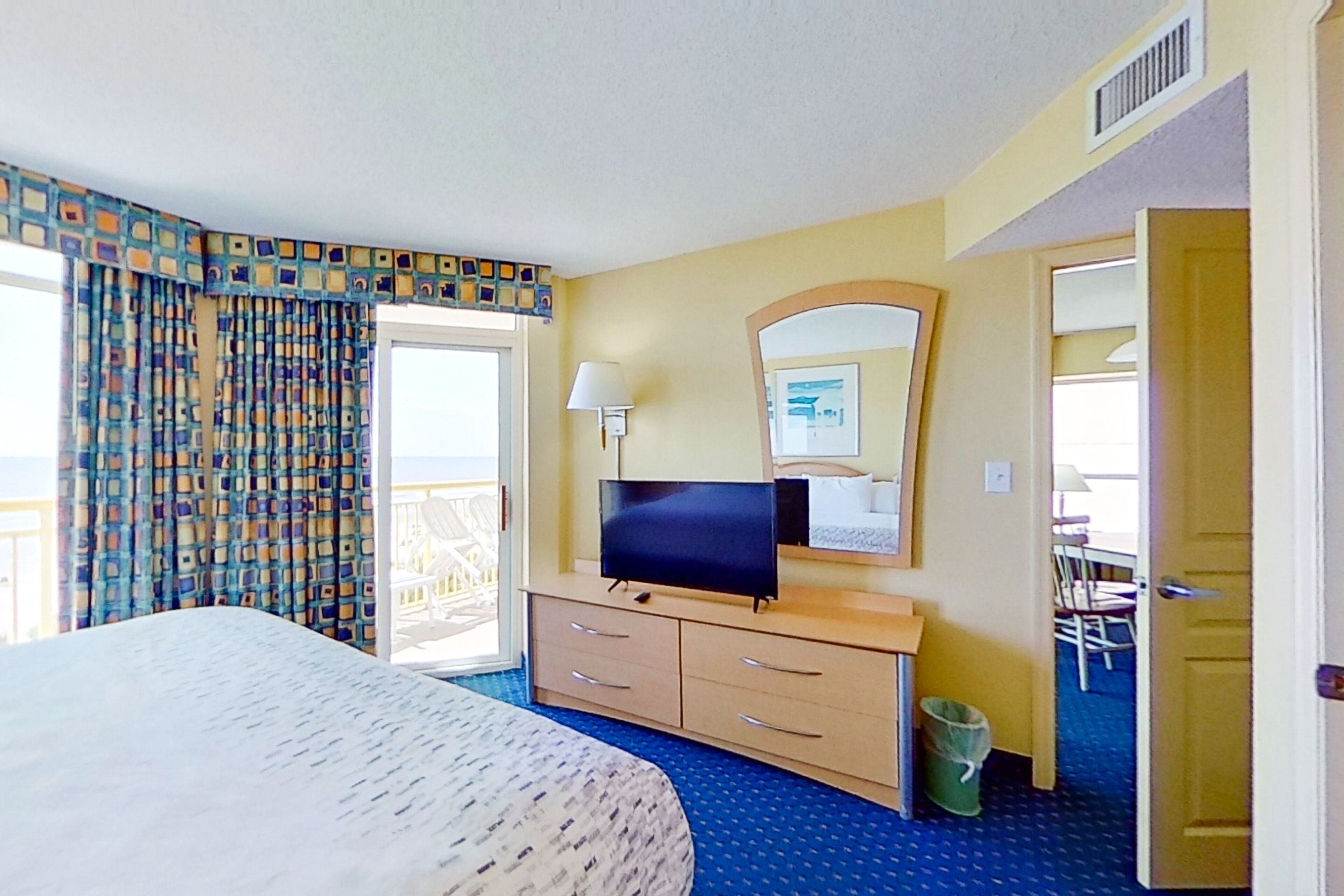 A hotel room with a bed, a television, a desk with drawers, a mirror, and a view of the balcony.