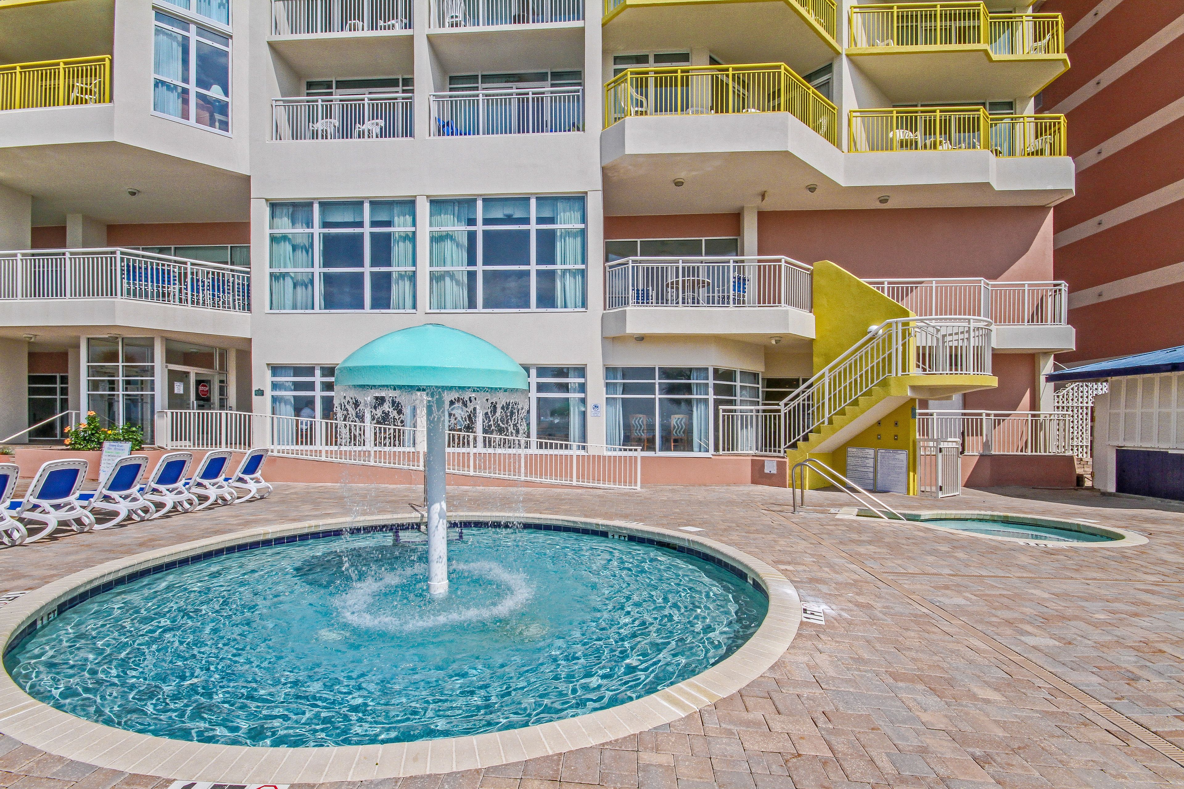 Une piscine avec des chaises longues, une fontaine et un immeuble à plusieurs étages doté de balcons.