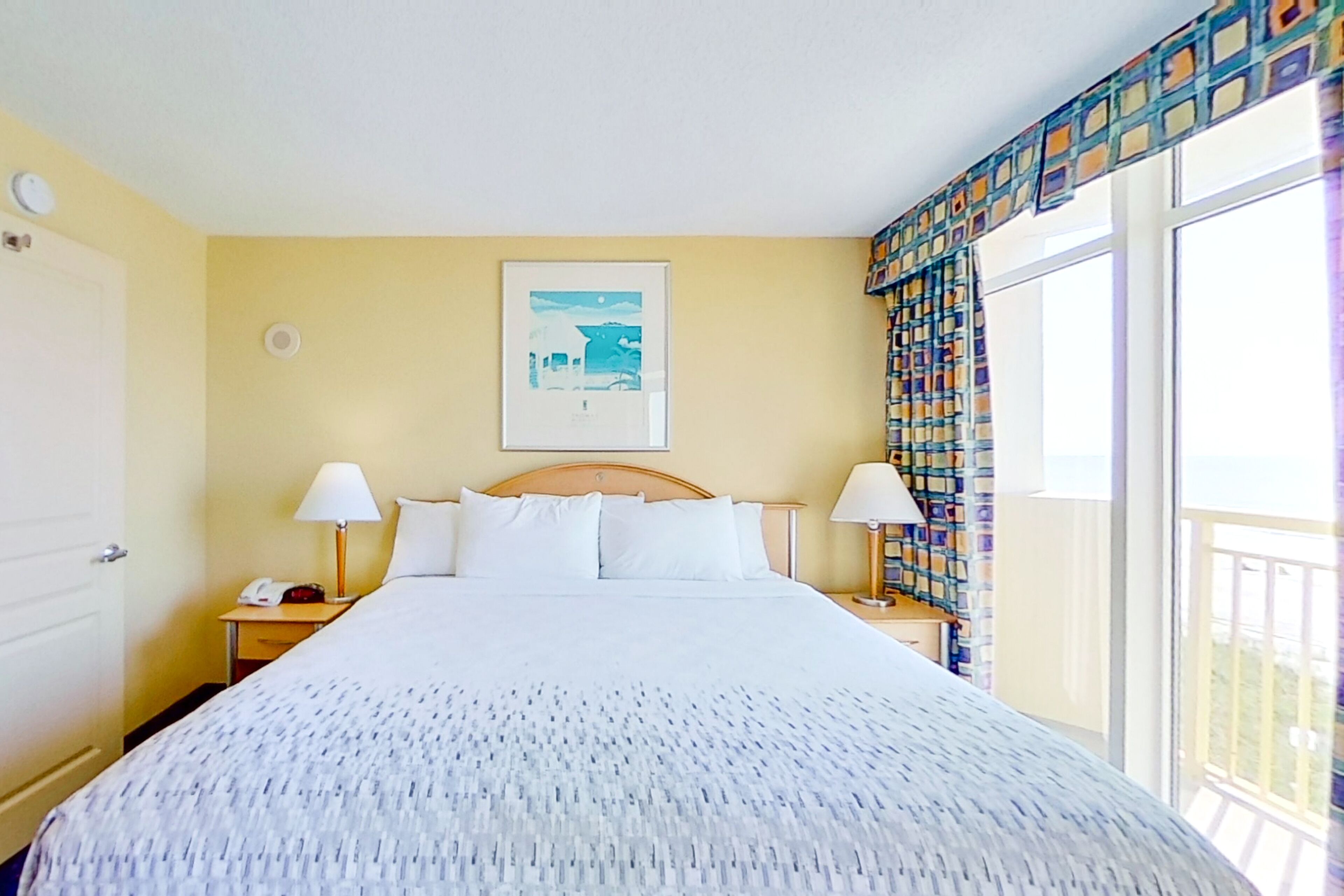 A hotel room with a large bed, two bedside tables with lamps, a framed picture on the wall, and a balcony with patterned curtains.