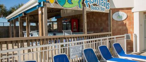 A beach bar named 'Fishtails' with a sign featuring a fish and a drink. There are lounge chairs in front of the bar.