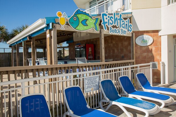 A beach bar named 'Fishtails' with a sign featuring a fish and a drink. There are lounge chairs in front of the bar.