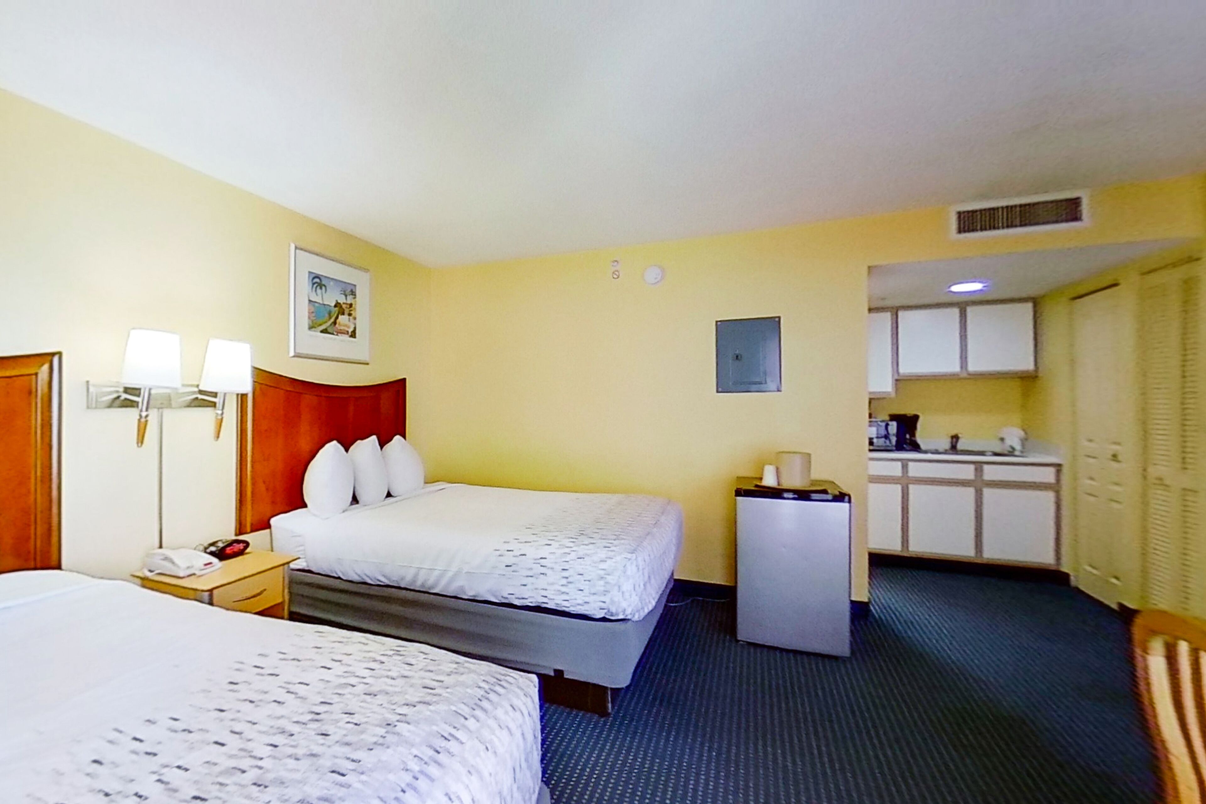 A hotel room with two beds, a desk with a lamp, a refrigerator, and a kitchenette.