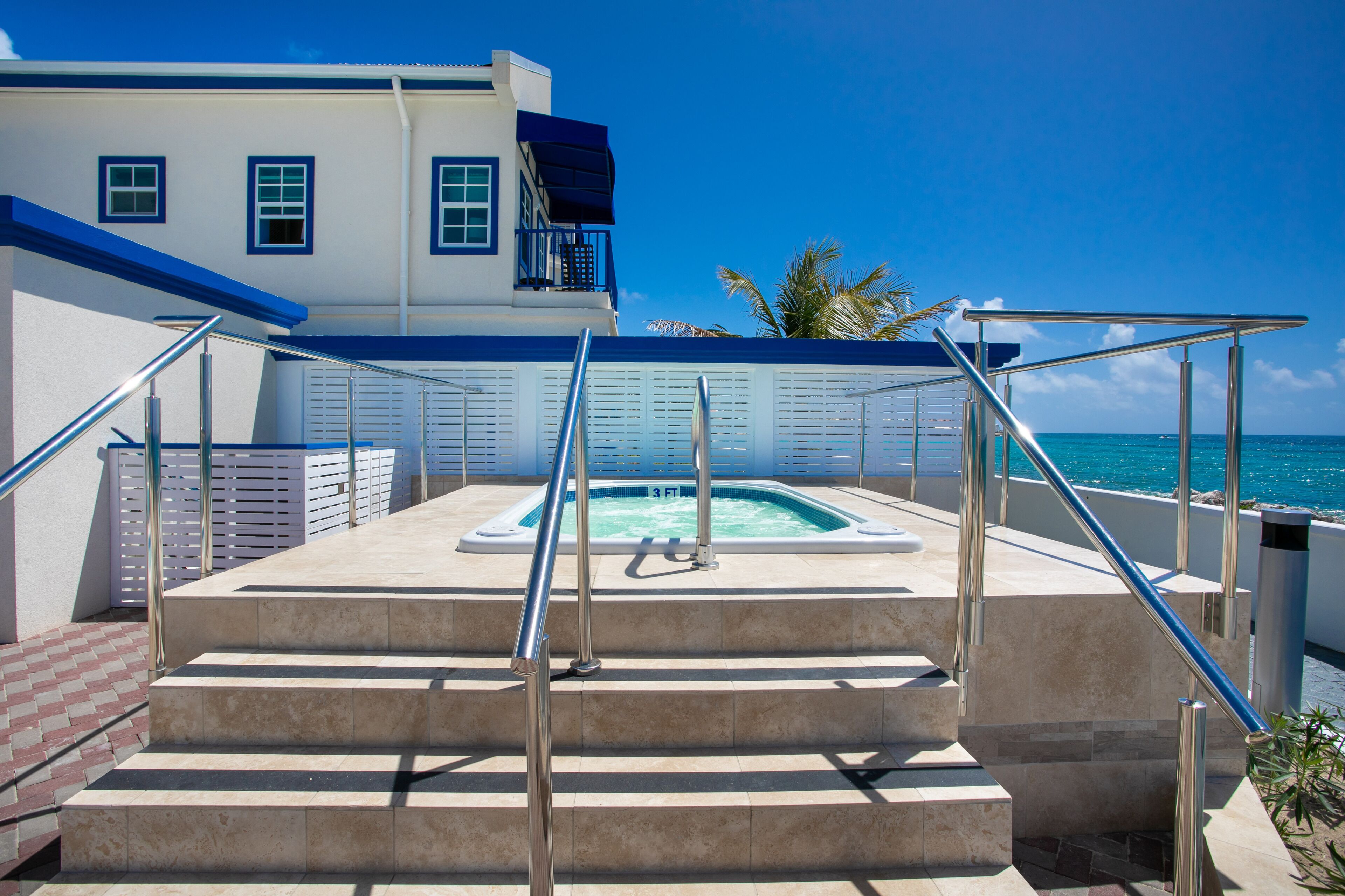 A pool area with steps leading up to a hot tub, surrounded by a white building with blue accents and a view of the ocean.