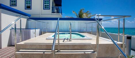 A pool area with steps leading up to a hot tub, surrounded by a white building with blue accents and a view of the ocean.