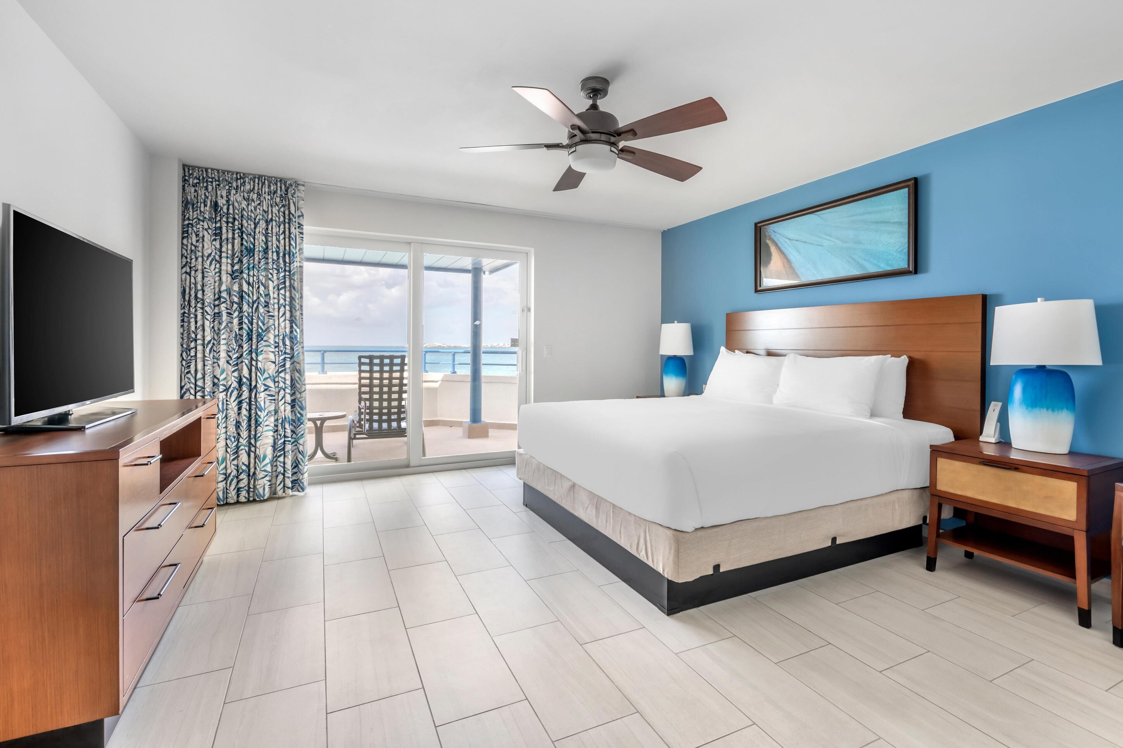 A hotel room with a bed, a TV, a ceiling fan, a nightstand, and a view of the ocean.