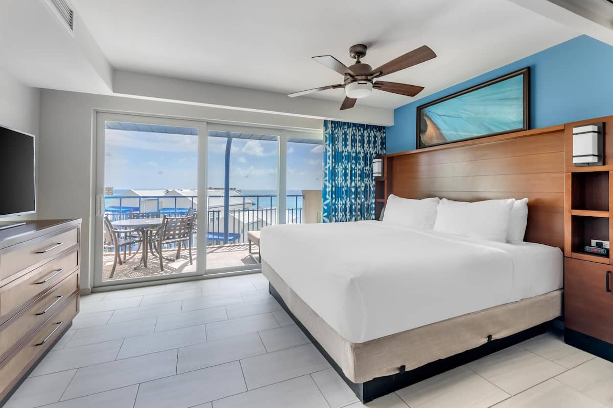 A hotel room with a large bed, a ceiling fan, a TV, a wooden headboard, a sliding glass door to a balcony, and a view of the sea.