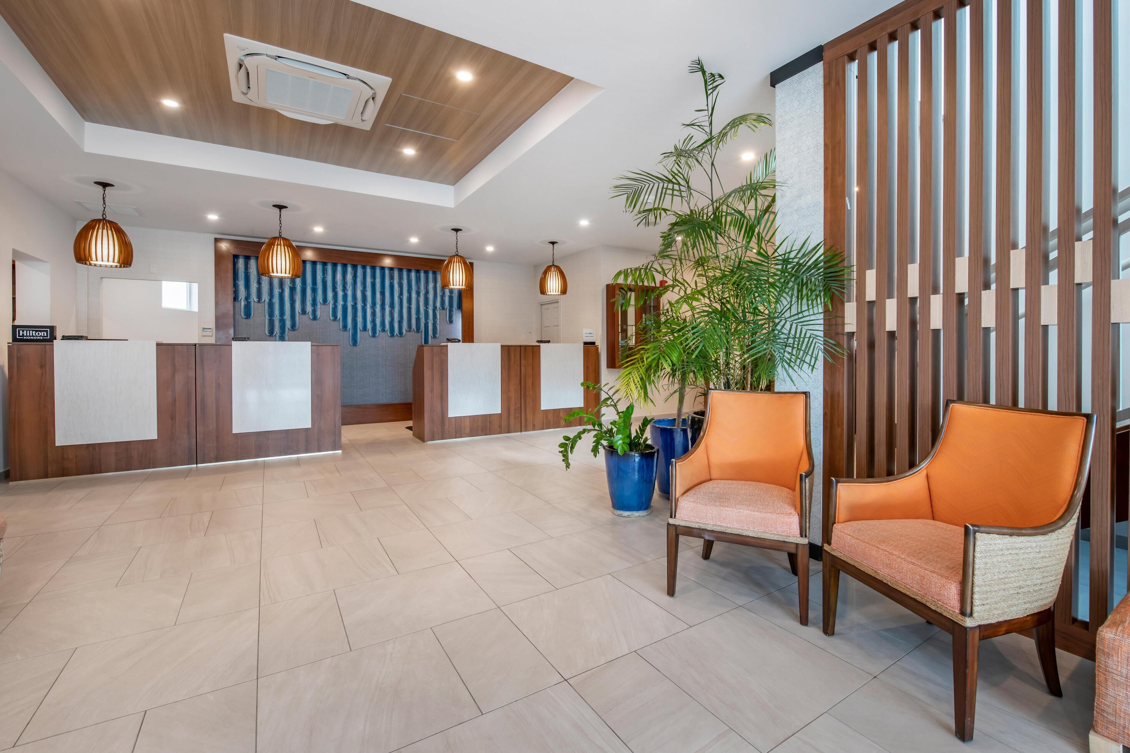 Zona de recepción de un hotel con un mostrador de madera, dos recepcionistas y un área de asientos con dos sillas y una planta en maceta.