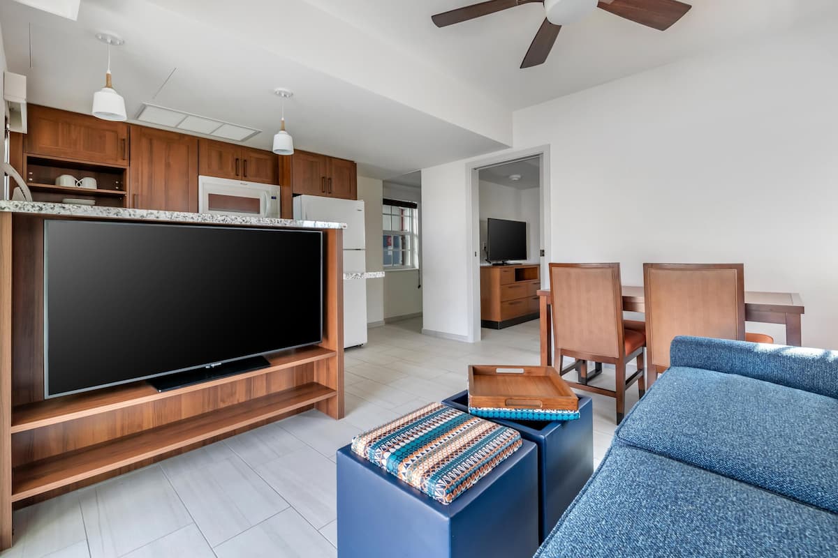 A modern living room with a flat-screen TV, wooden cabinets, a dining area with chairs, and a ceiling fan.