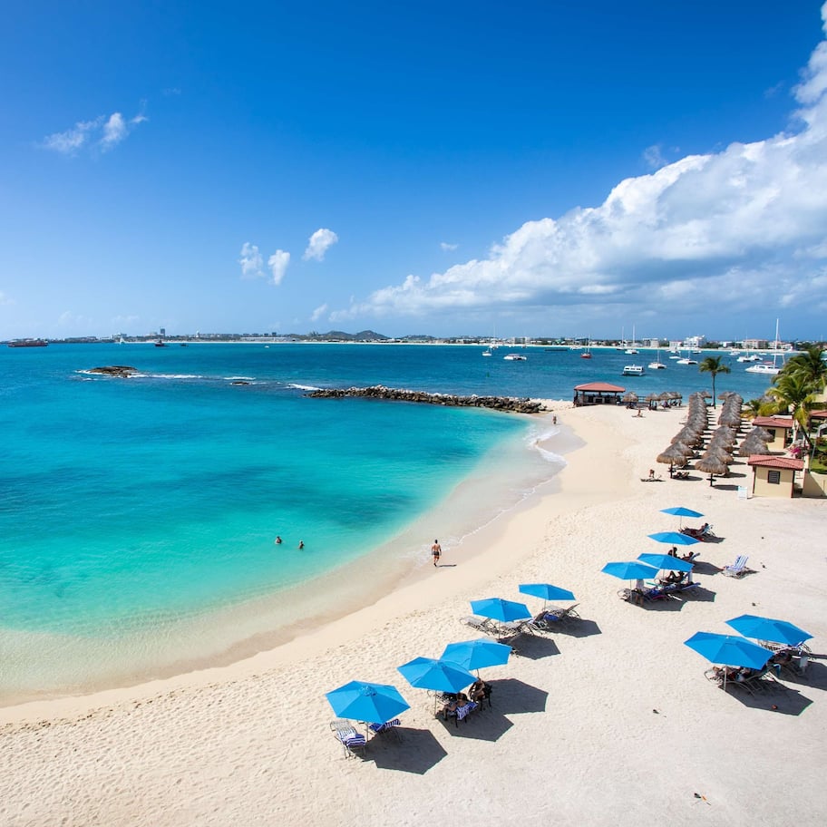 A beach with clear turquoise waters, a sandy shore, and several blue umbrellas.