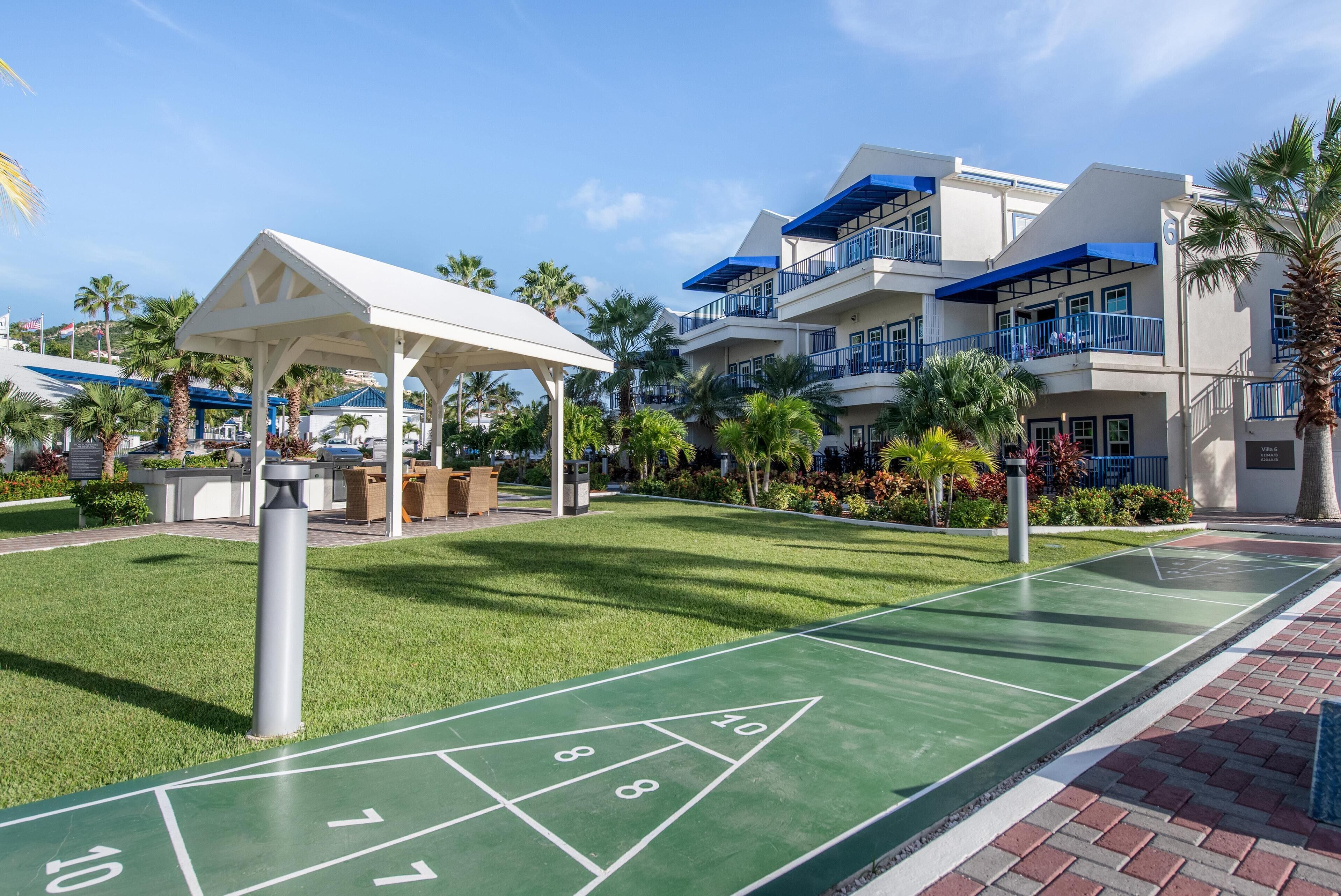 A shuffleboard court, a covered seating area, and a building with balconies and a blue awning.