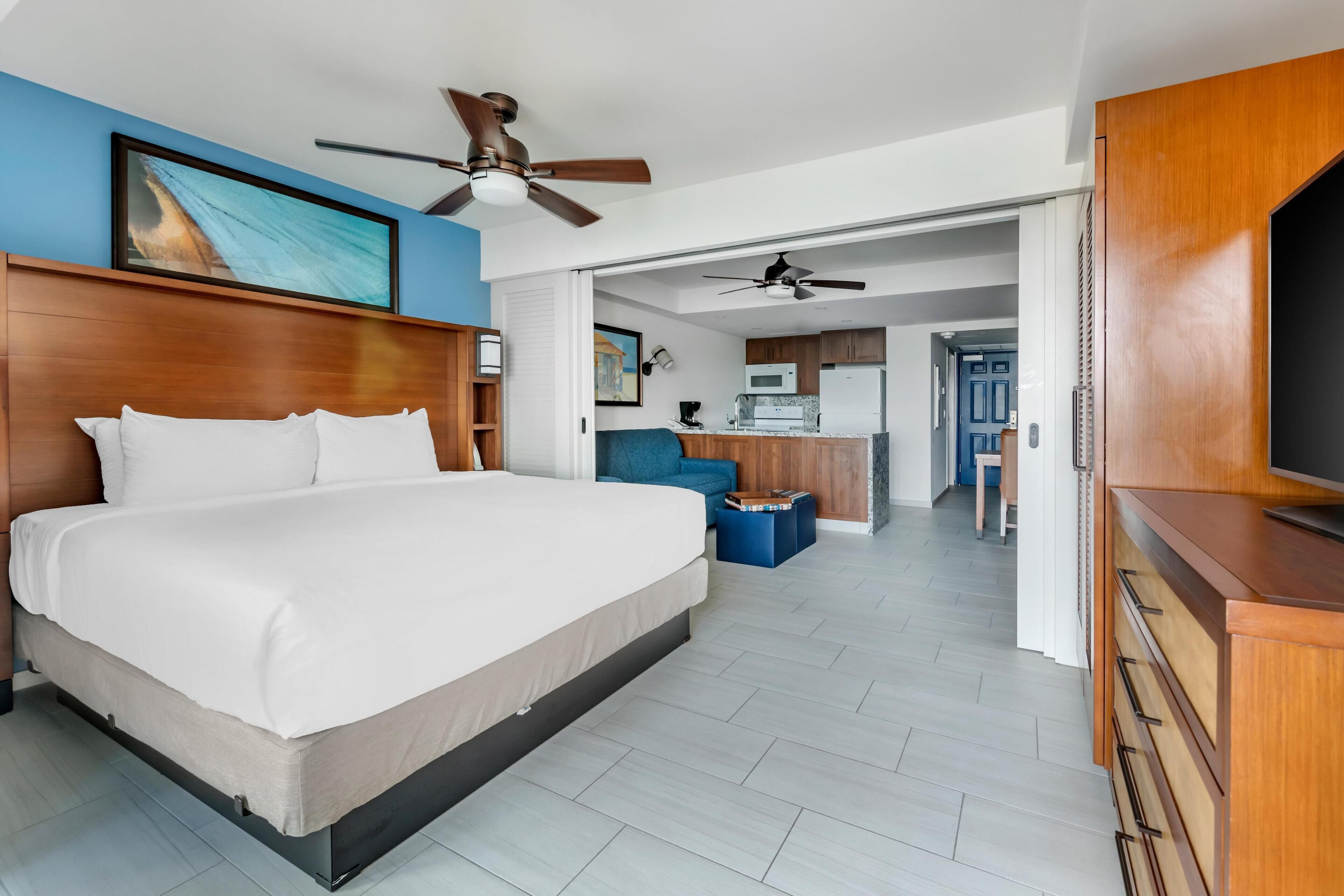 A modern hotel room with a large bed, a ceiling fan, a kitchenette, and a TV.