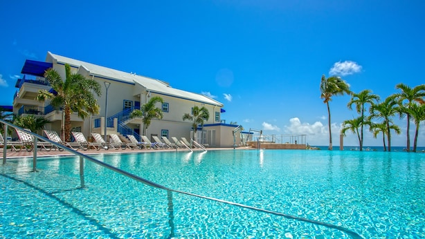 Une piscine à l’eau d’un bleu cristallin, un bâtiment doté de balcons et des palmiers.