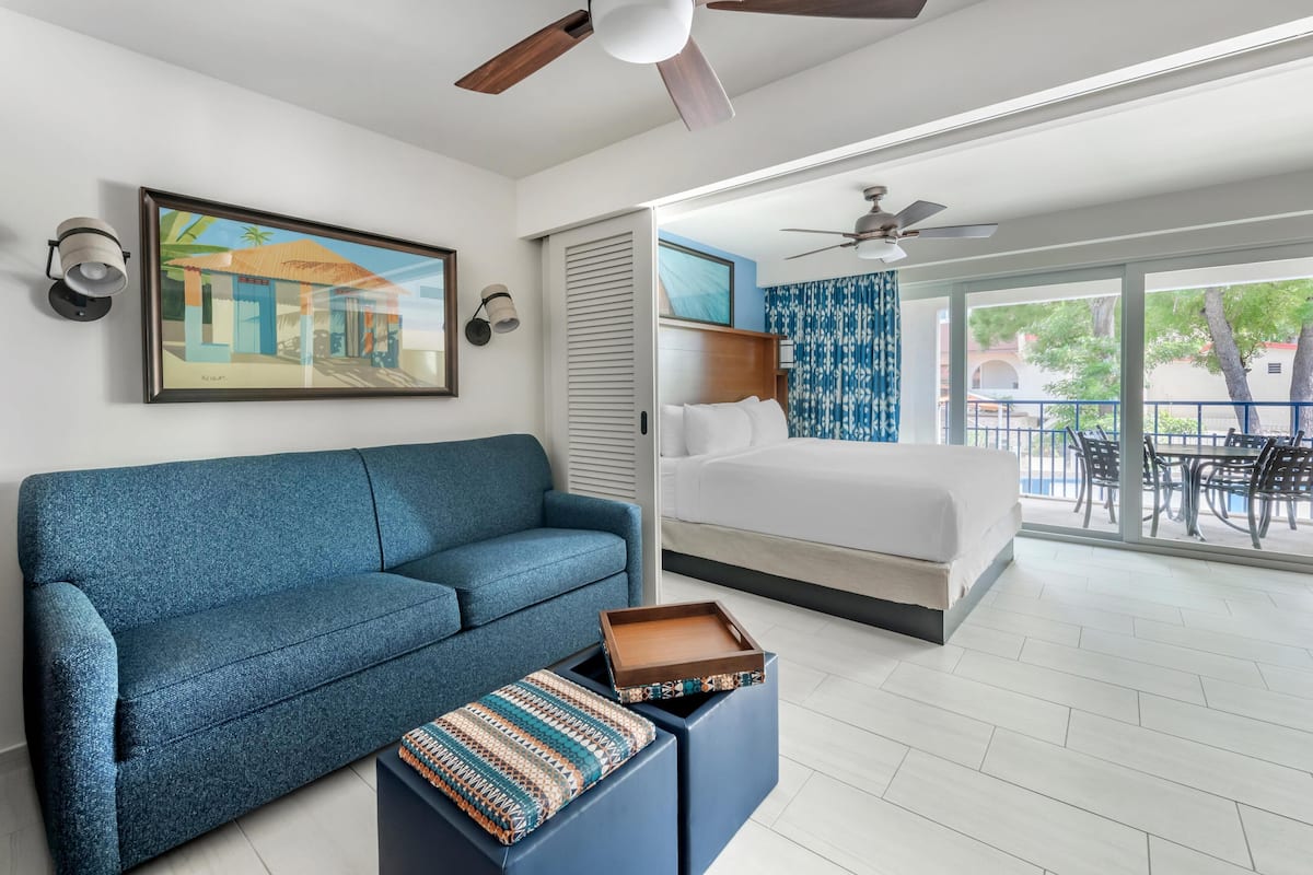 A modern hotel room with a blue sofa, a bed, a balcony with a view, and a ceiling fan.