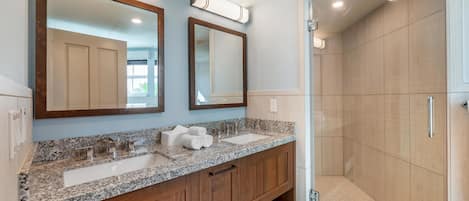 A bathroom with a double sink vanity, wooden cabinets, a large mirror, and a walk-in shower.
