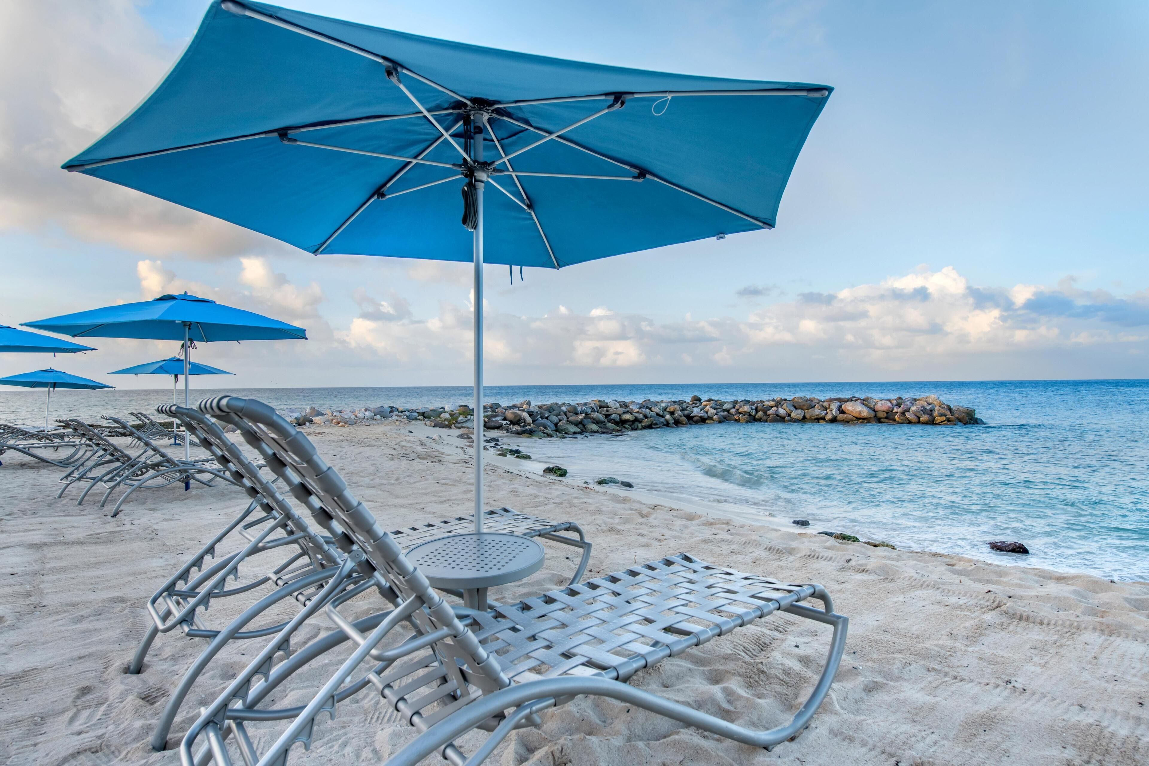 A beach with metal lounge chairs and blue umbrellas, a rocky shoreline, and calm sea.