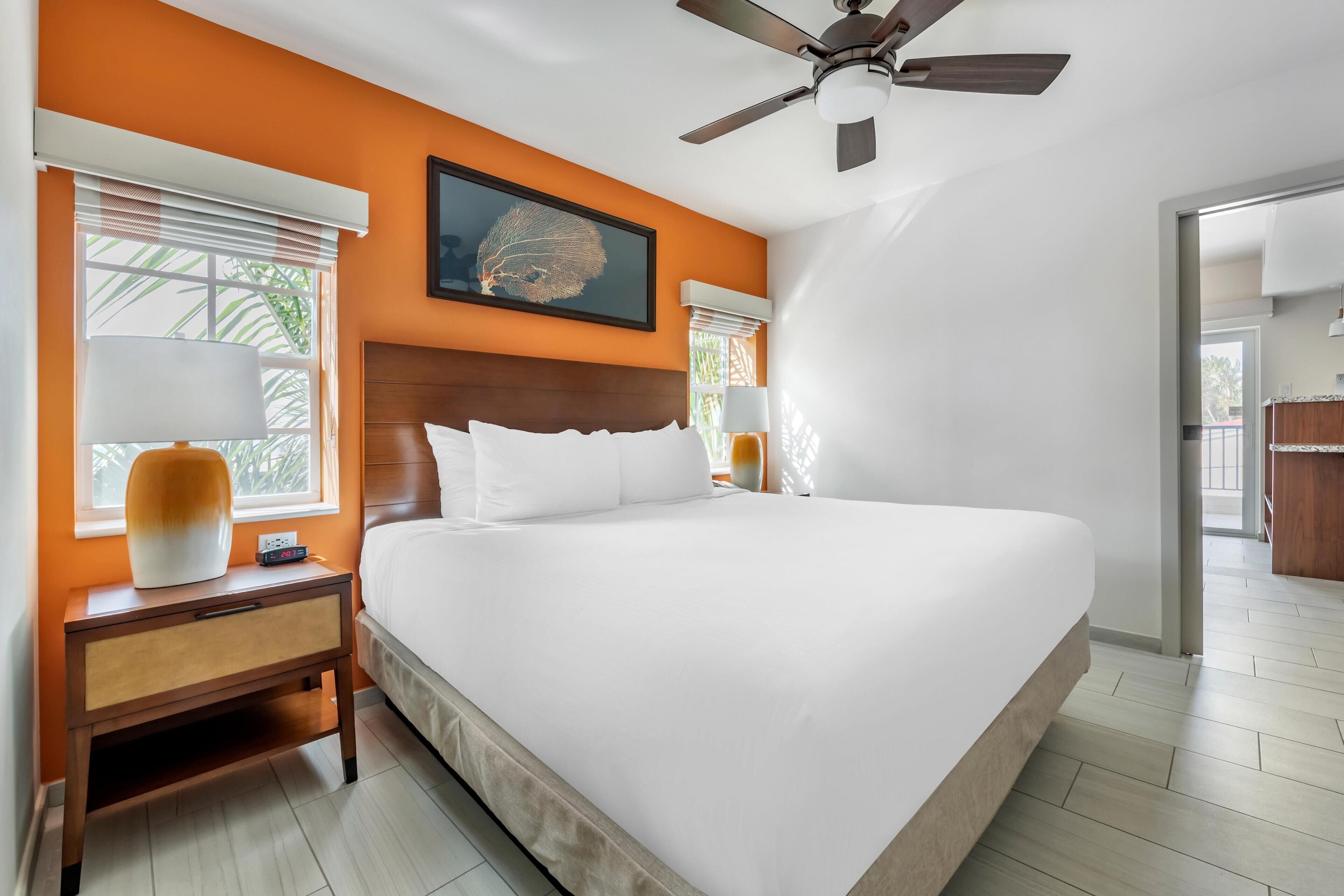 A large bed with white bedding, a wooden nightstand with a lamp, and a ceiling fan in a room with orange walls and a large window.
