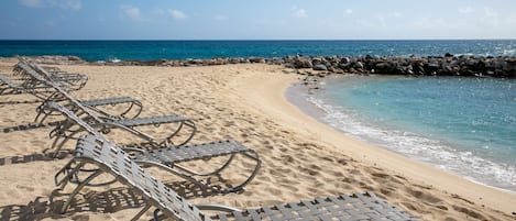 A beach with metal lounge chairs aligned along the shoreline, a rocky breakwater, and clear blue water.