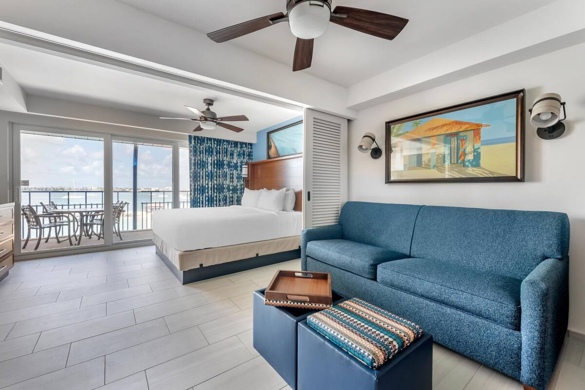 A modern hotel room with a bed, a blue sofa, a dining area with a table and chairs, and a balcony with a view of the sea.