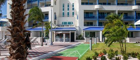 A hotel with a modern design, featuring a rooftop pool, a tennis court, and a landscaped garden with palm trees.