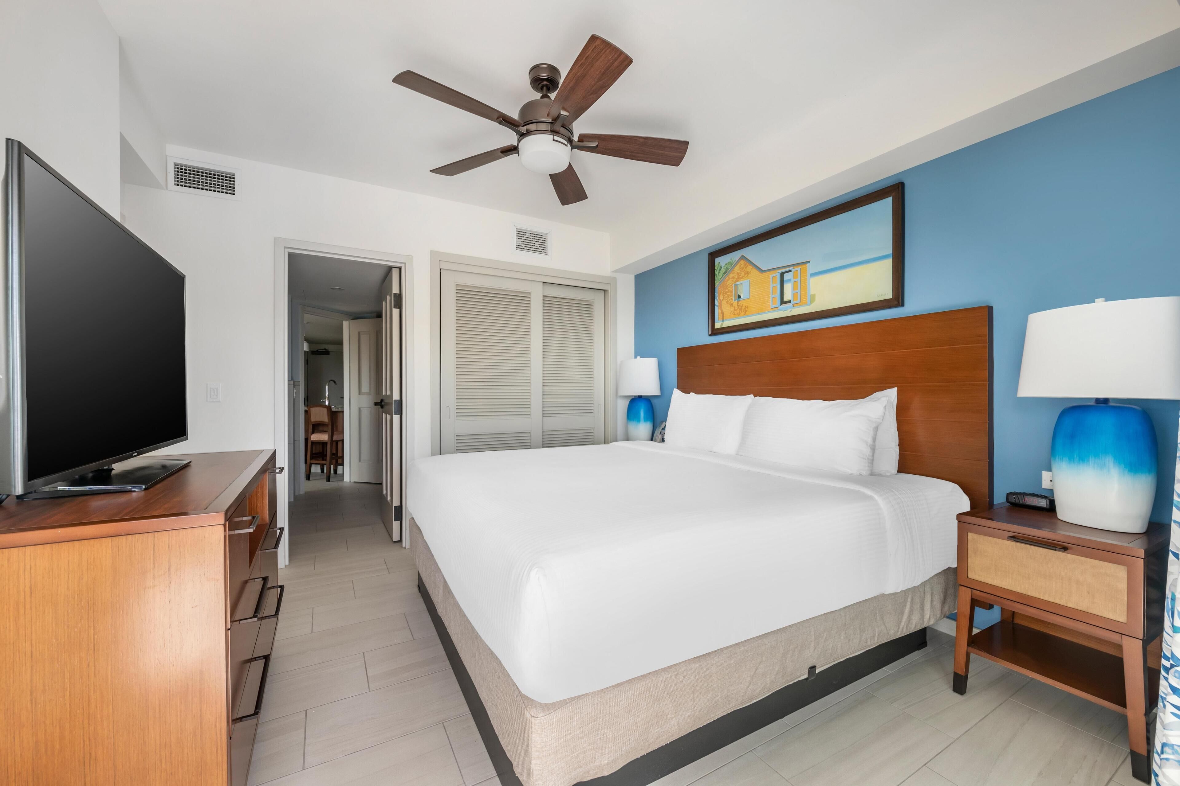 A hotel room with a large bed, a ceiling fan, a TV, and a painting on the wall.