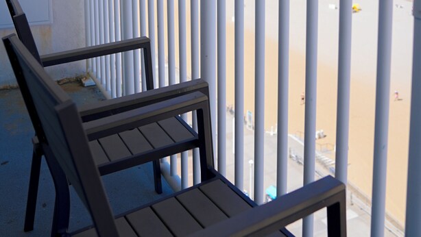 A balcony with two wooden chairs, overlooking a beach with a clear sky and calm sea. - Sun and Sand Resort Oceanfront Suites  (Virginia Beach)