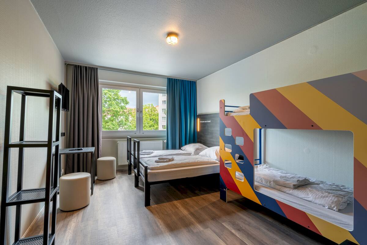 A dormitory room with a bunk bed, a desk, a chair, and a window with curtains.