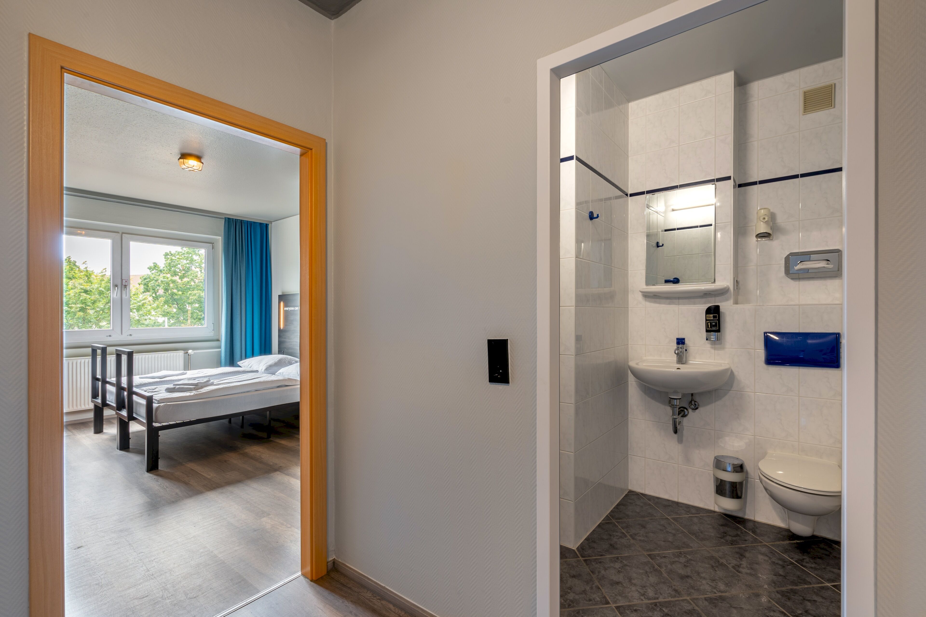 A modern hotel room with a wooden bed, a bathroom with a sink and toilet, and a view of the outdoors through a window.