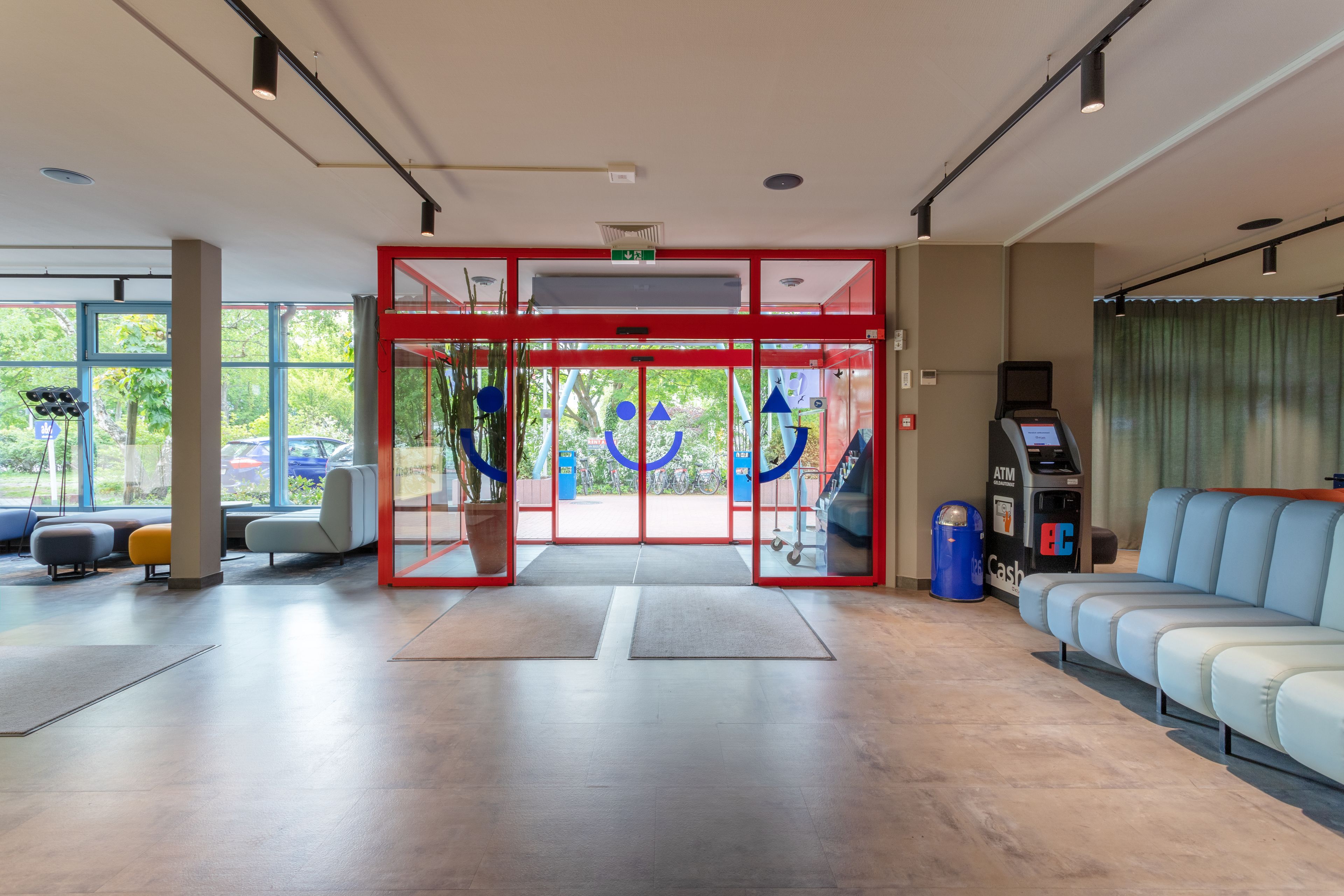 Un vestíbulo de hotel moderno con entrada roja, zona de estar y un cajero automático.