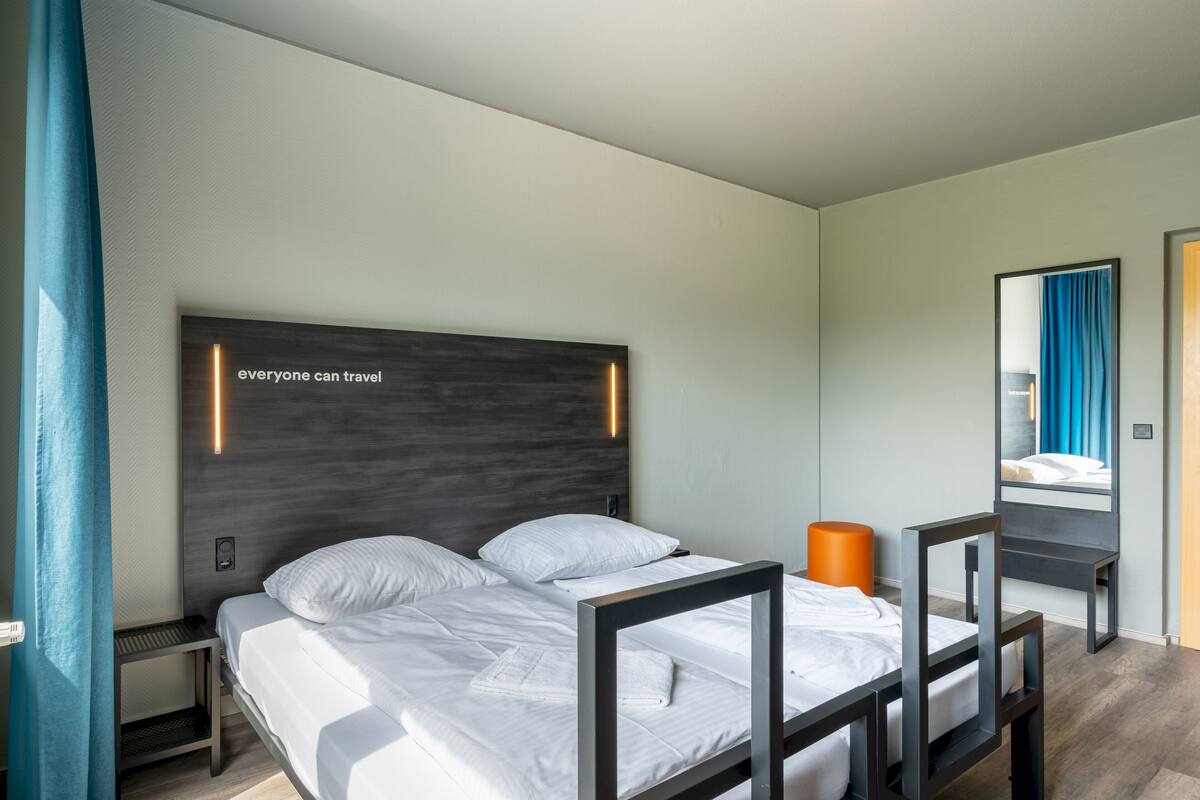A modern hotel room with a large bed, a bedside table, a nightstand, a mirror, and a view of the city through the window.