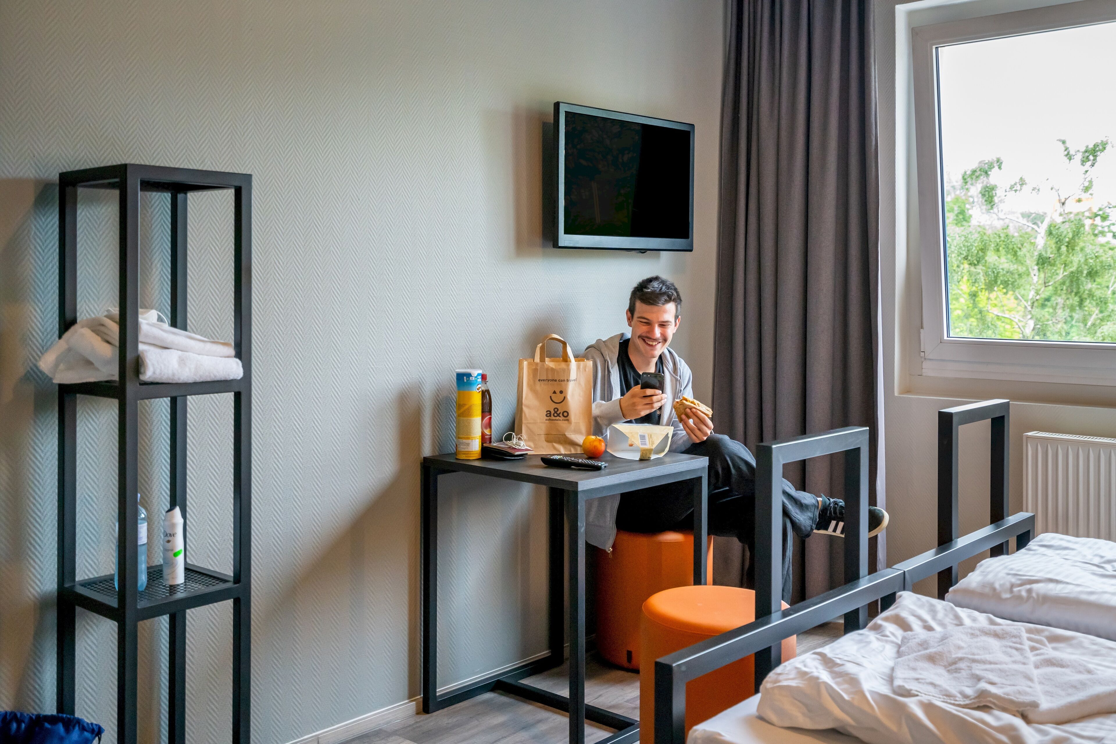 Una camera d'albergo con un letto, un tavolino con una sedia, una TV a parete e una finestra vista alberi.