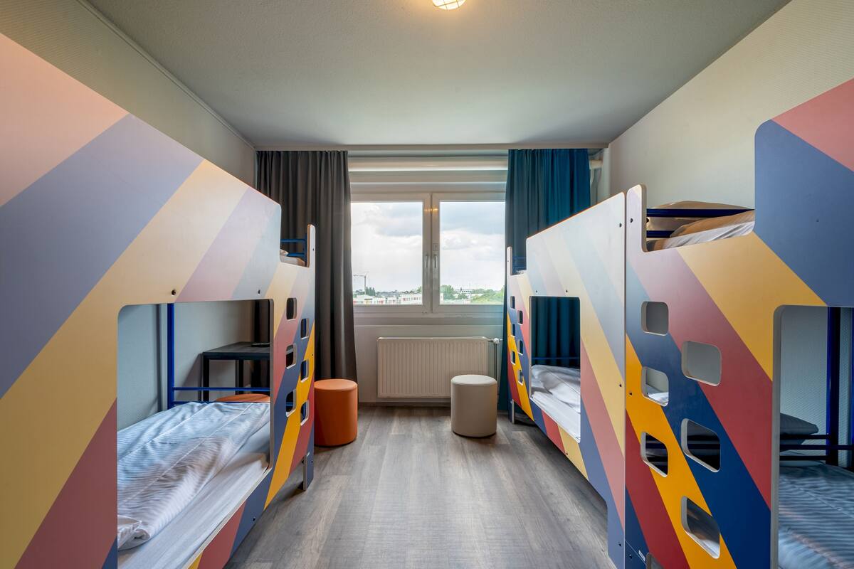 A hotel room with bunk beds, a small table, a chair, a window with a view, and a radiator.