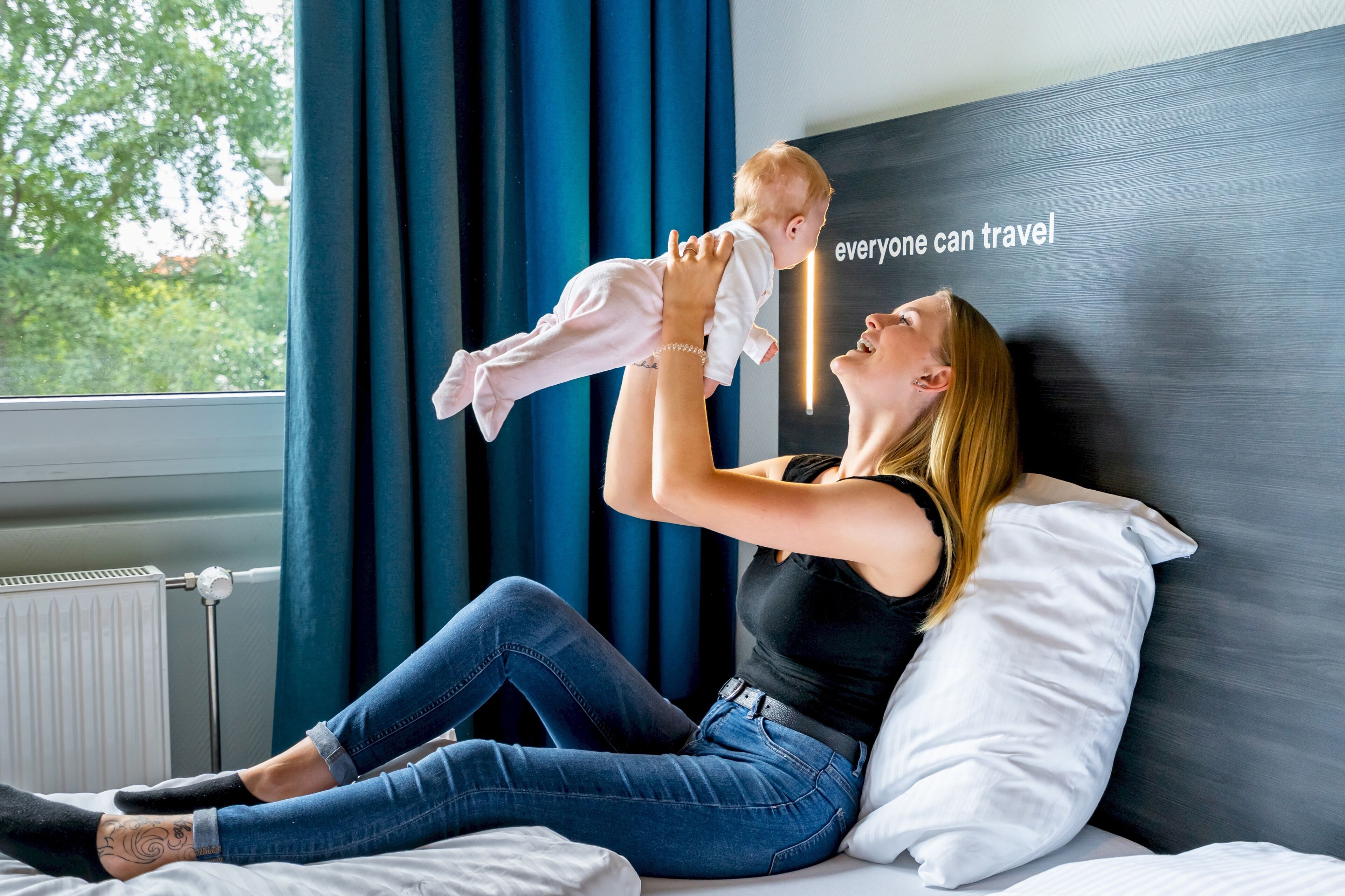 Une femme est assise sur un lit avec un enfant, et elle le tient en l’air.