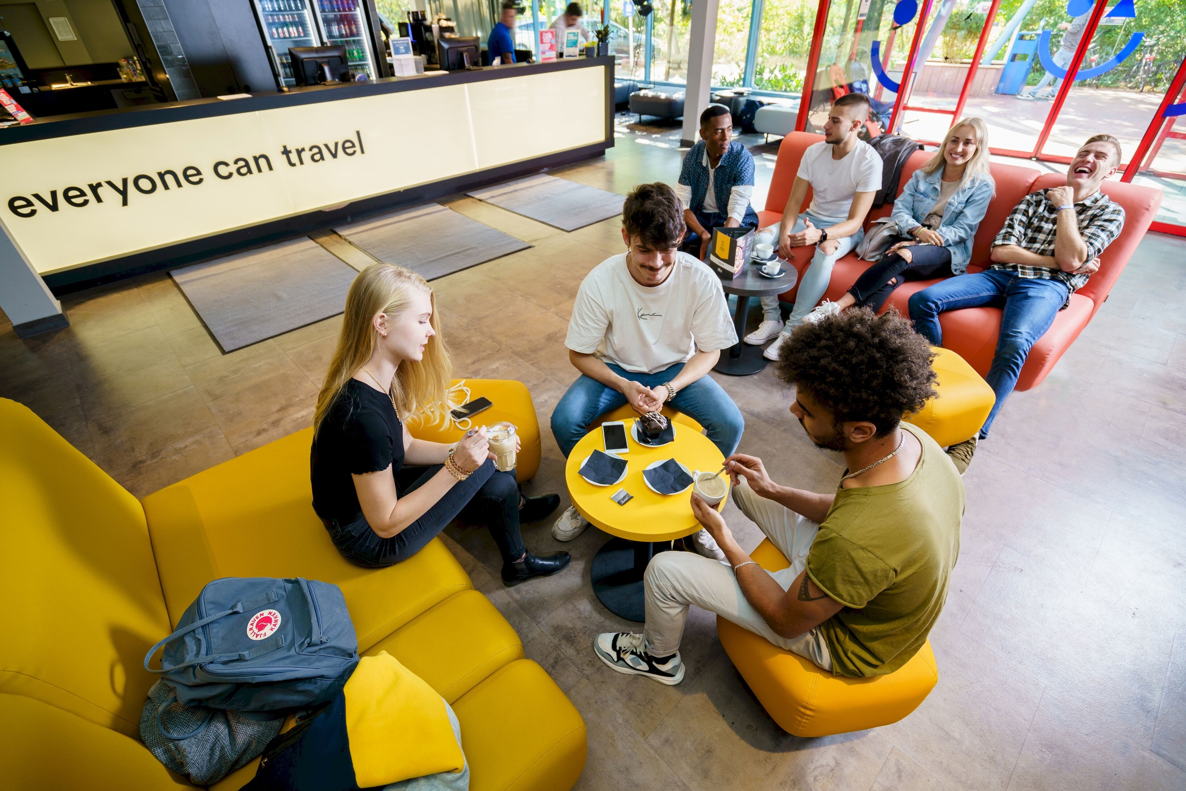 A group of people sitting on yellow and orange seating areas in a modern indoor space with a reception desk in the background.