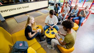 A group of people sitting on yellow and orange seating areas in a modern indoor space with a reception desk in the background.
