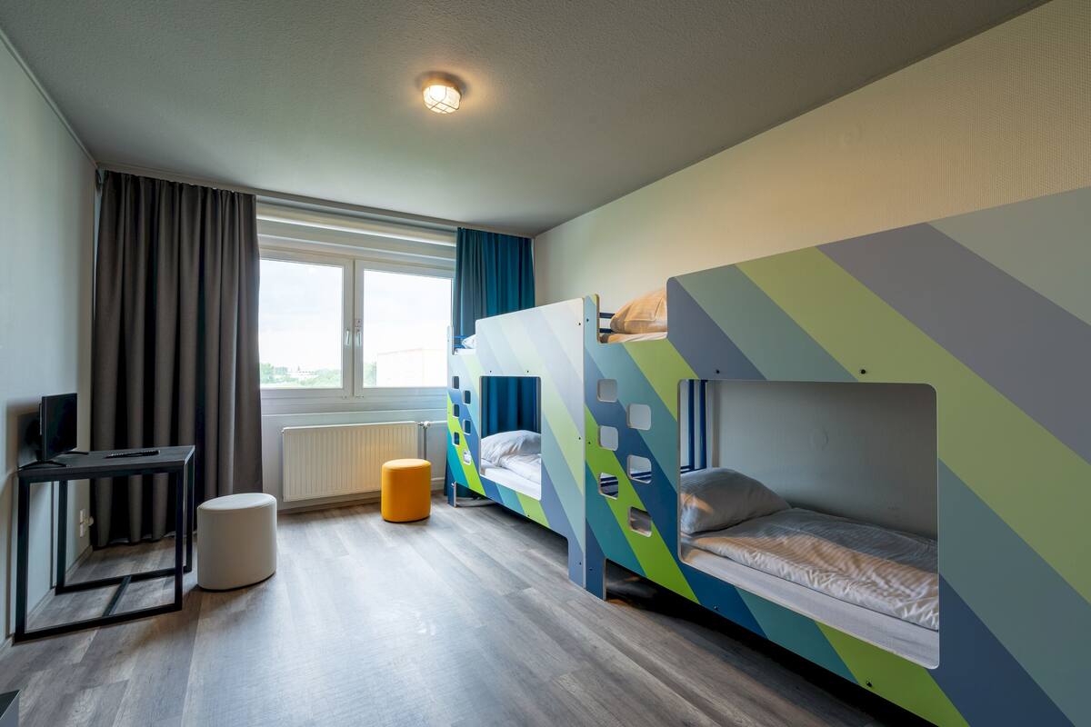 A modern hotel room with a wooden floor, a desk with a chair, a television, and two bunk beds with colorful geometric patterns.