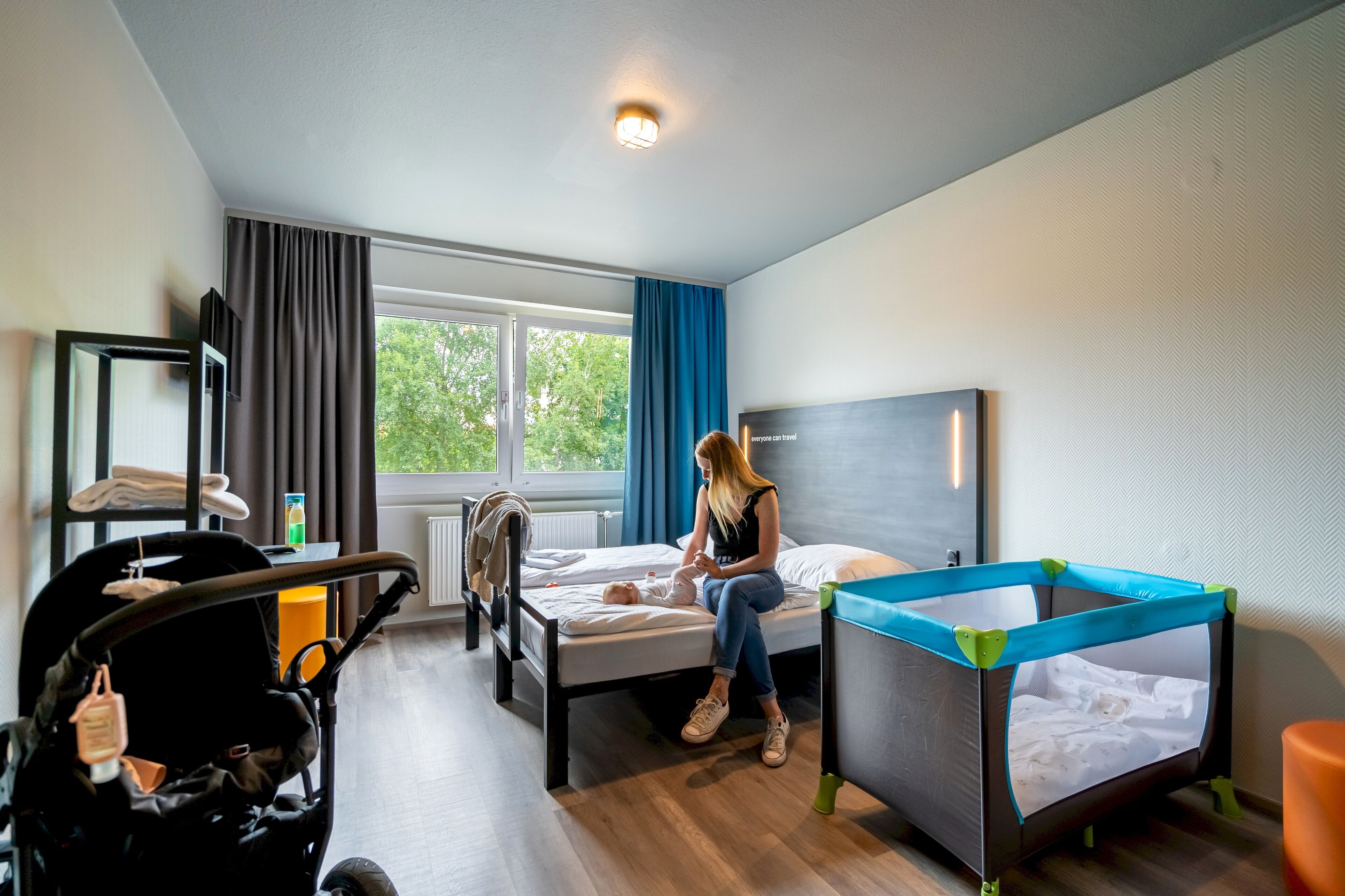 Habitació d'hotel amb llit, bressol, cotxet i finestra amb cortines.