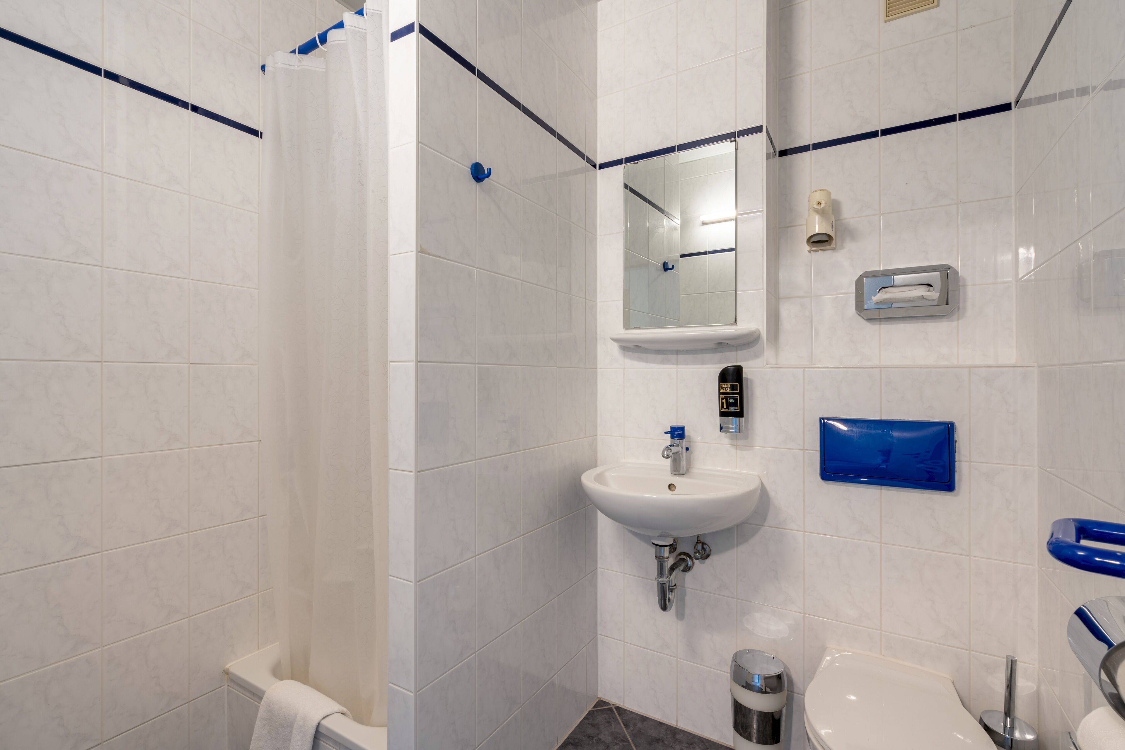 Un baño moderno con azulejos blancos, un lavamanos blanco, un espejo y un perchero azul.