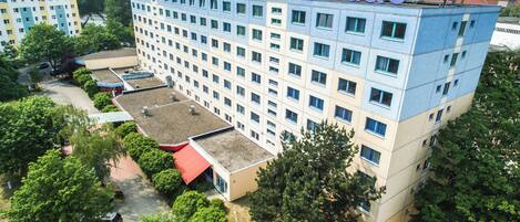 A multi-story hotel with a rooftop terrace, surrounded by greenery and other buildings.