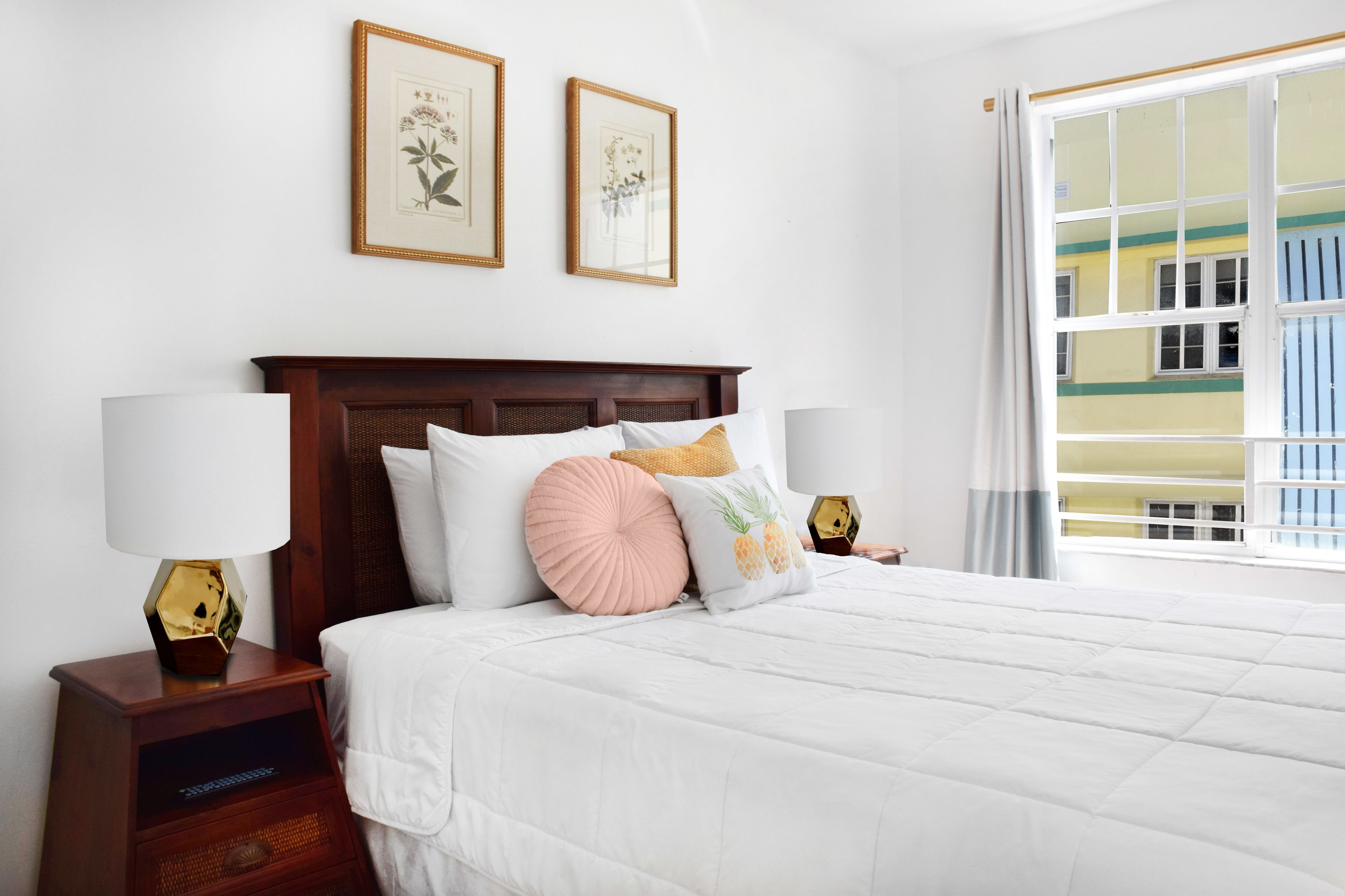 A neatly made bed with a white comforter and multiple pillows, including a pineapple design. There are two wall-mounted framed botanical prints above the bed.