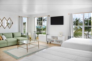 Habitación de hotel moderna con sofá verde, una mesa de centro de vidrio, cama blanca y un ventanal con vista a palmeras.