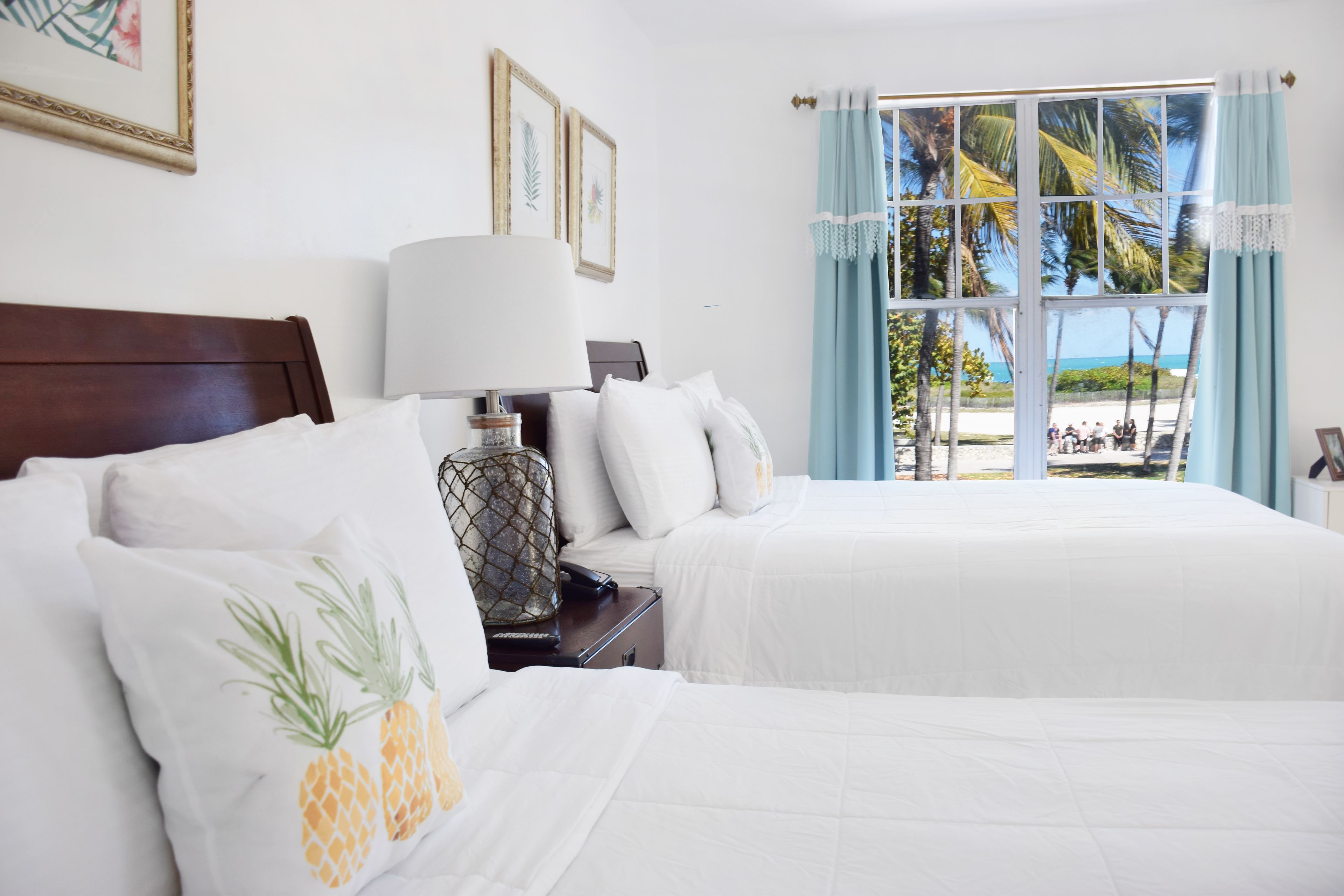 Een hotelkamer met twee bedden, witte beddengoed, een houten hoofdbord, een nachtkastje met een lamp en uitzicht op een strand met palmbomen.